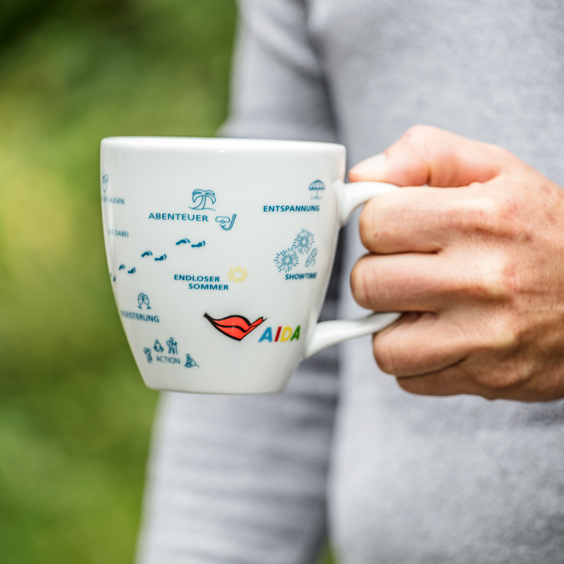 Weiße Keramiktasse mit Henkel, dekoriert mit bunten maritimen Urlaubsmotiven – Kreuzfahrtschiff-Silhouette, stilisierte Weltkarte mit Reiserouten, Wellen, Sonne und Palmen – mit dezentem ‚AIDA‘-Logo unter dem Rand